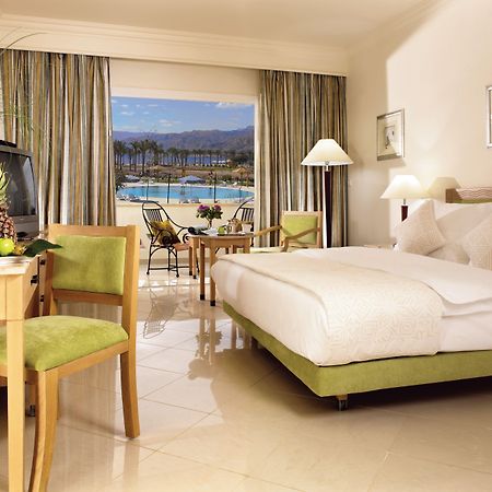 Movenpick Taba Resort & Spa Værelse billede The photo shows an elegant hotel room featuring a large bed with white linens and several pillows. There is a small dining table with green chairs beside it and a television on a wooden unit. Large windows with sheer curtains let in natural light, of