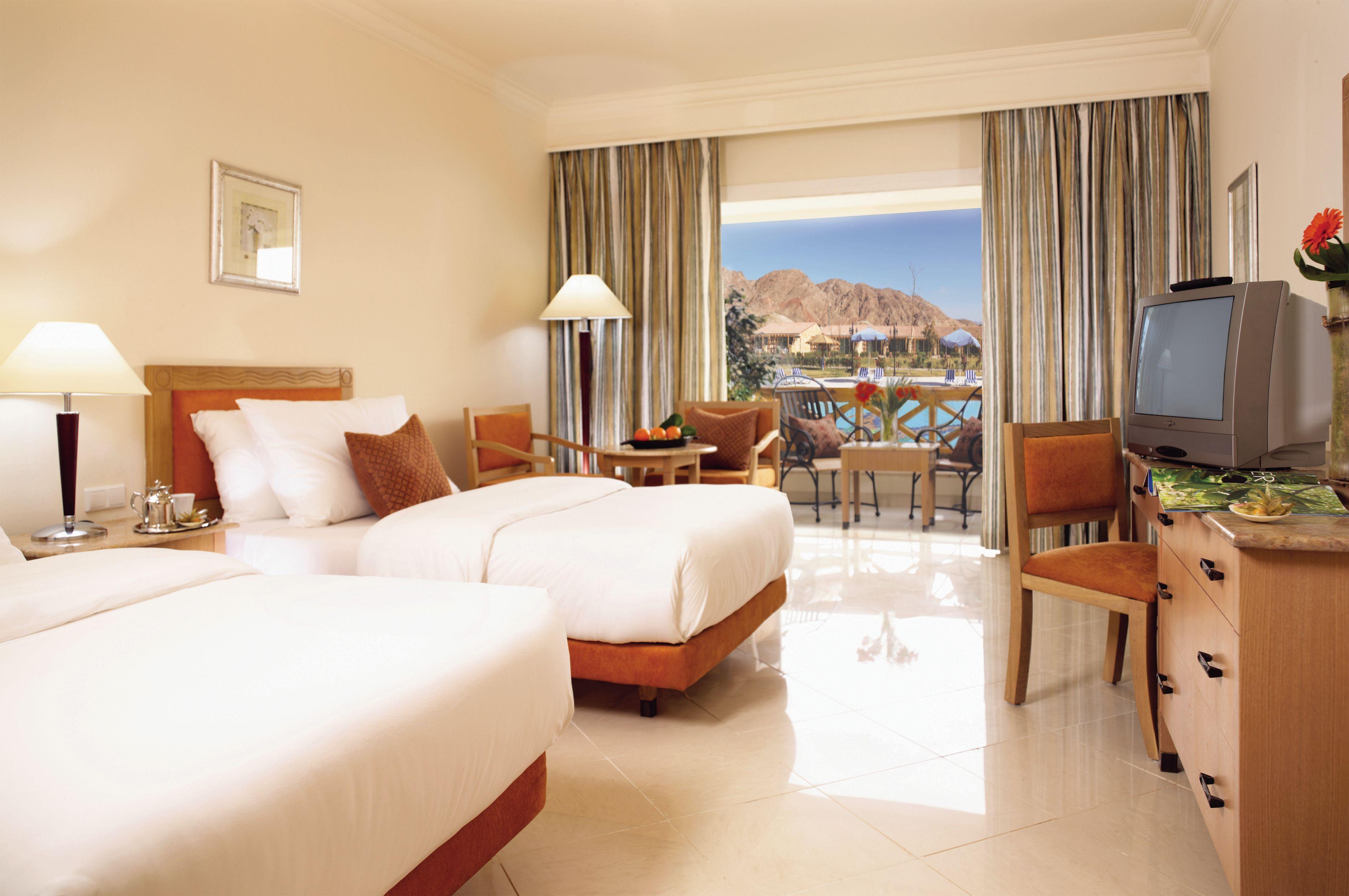 Movenpick Taba Resort & Spa Værelse billede The photo shows a hotel room with two twin beds placed side by side. The bedding is light in color, and there are decorative pillows on each bed. A wooden bedside table with a lamp is positioned next to one of the beds. 

In the background, there are