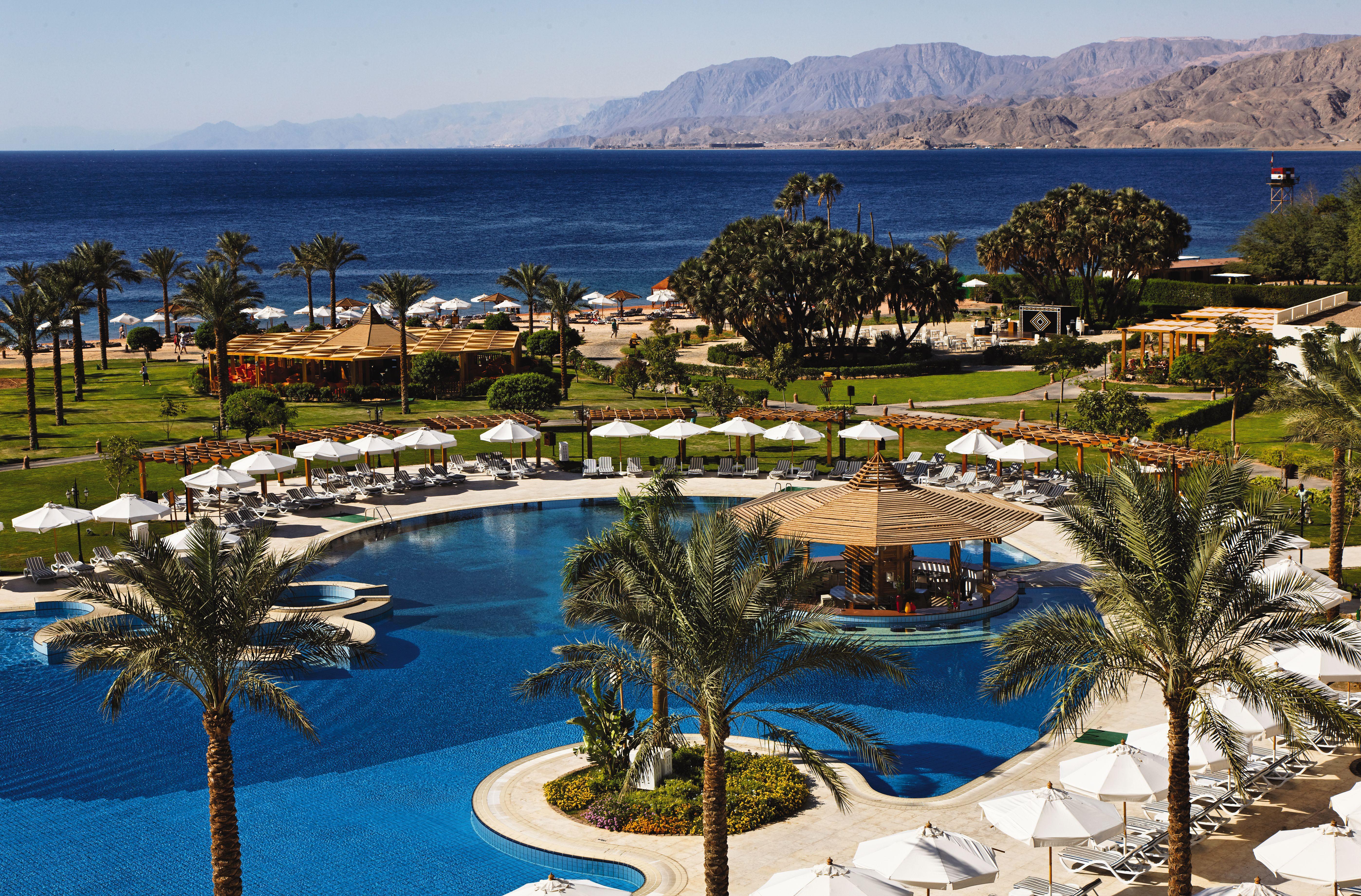 Movenpick Taba Resort & Spa Eksteriør billede The photo shows a picturesque resort setting by the sea. In the foreground, there is a beautifully designed swimming pool with curves and a central gazebo or shade structure. Surrounding the pool are lounge chairs and umbrellas, suggesting a relaxing
