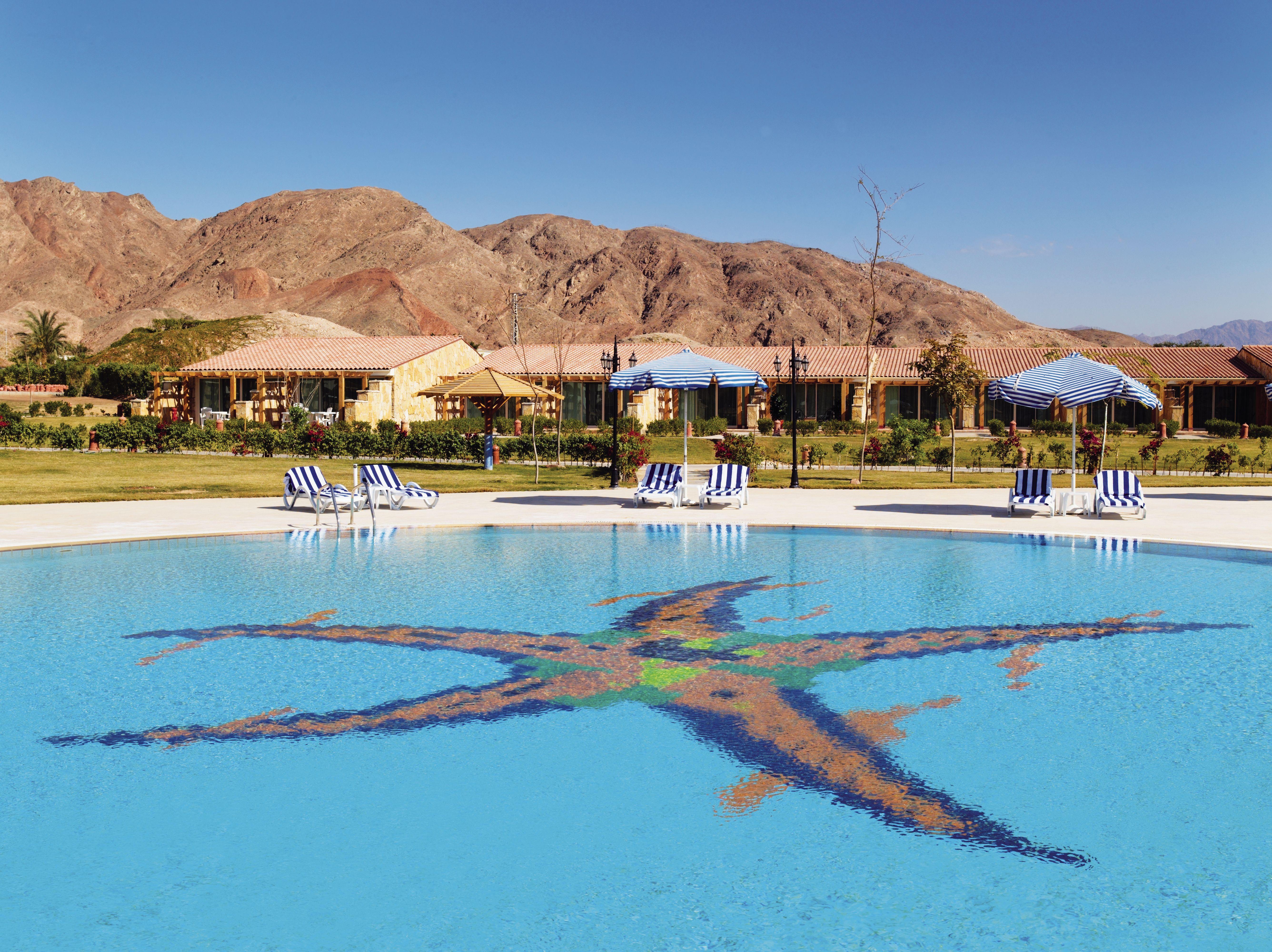 Movenpick Taba Resort & Spa Eksteriør billede The photo shows a swimming pool with a colorful star design at the bottom. There are several lounge chairs arranged around the pool, and a few umbrellas for shade. In the background, there are rolling hills or mountains, and a few buildings, likely a