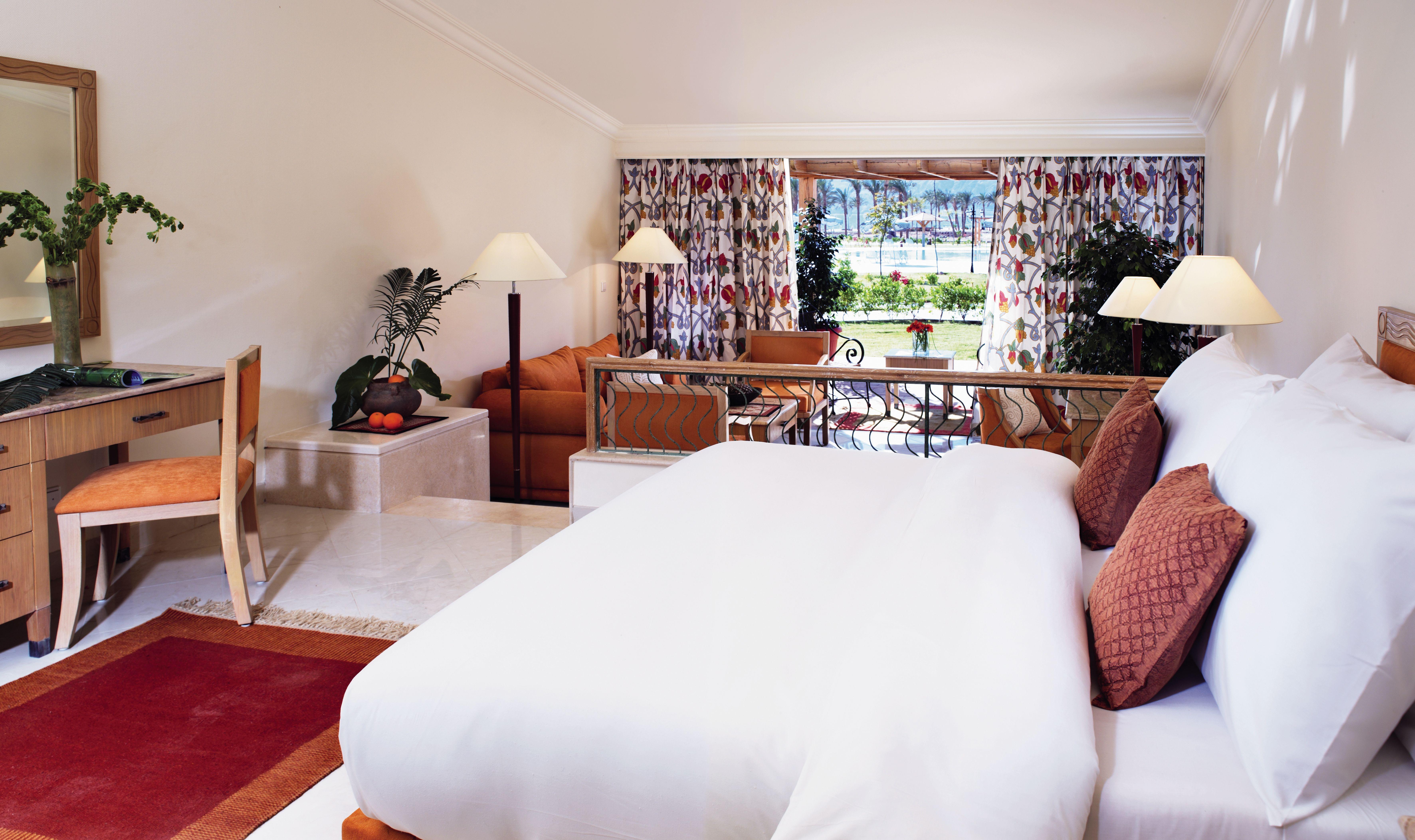 Movenpick Taba Resort & Spa Eksteriør billede The photo shows a spacious and elegantly designed hotel room or suite. In the foreground, there's a large bed with white bedding, complemented by decorative cushions. To the side, there’s a desk and chair, along with a bowl of fruit. 

In the backgro