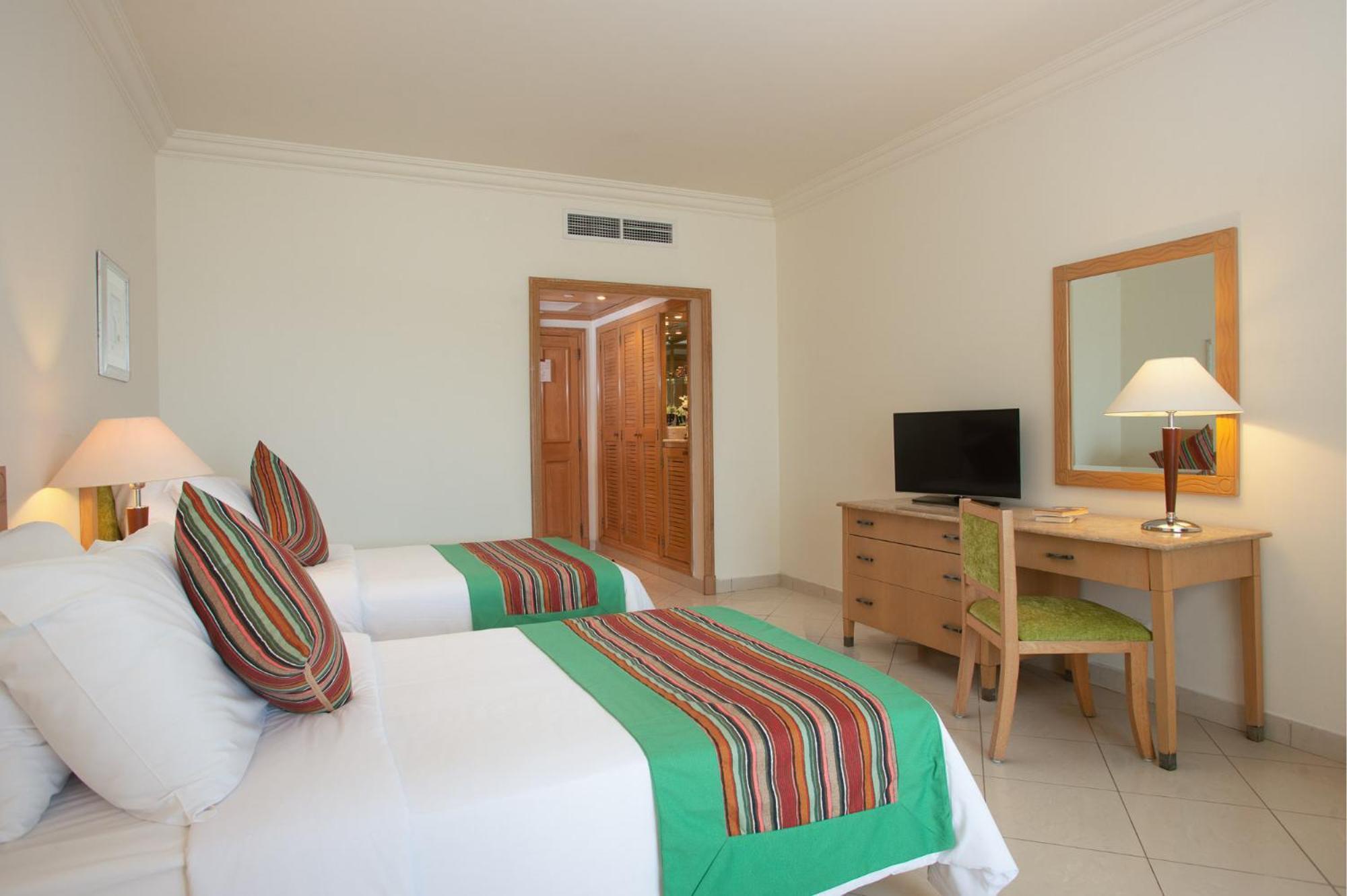 Movenpick Taba Resort & Spa Eksteriør billede The photo shows a hotel room featuring two beds with colorful striped throw blankets. There are decorative pillows on each bed, and the overall decor appears bright and modern. A wooden desk with a chair is positioned next to a TV, and there is a mir
