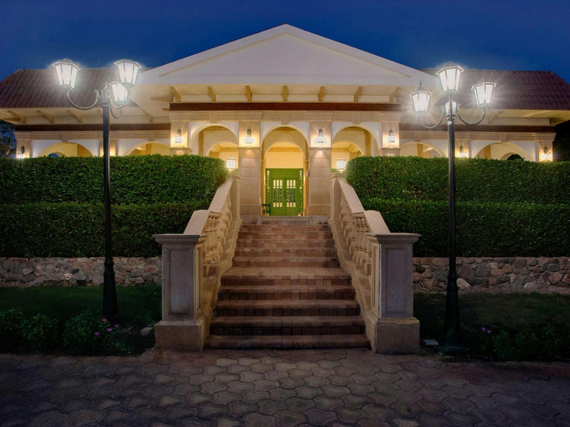 Movenpick Taba Resort & Spa Eksteriør billede The photo shows a large house or villa with a grand entrance. The structure features a sloped roof and is adorned with columns and detailed architectural elements. There are steps leading up to a double-door entry that is painted green. 

On either s