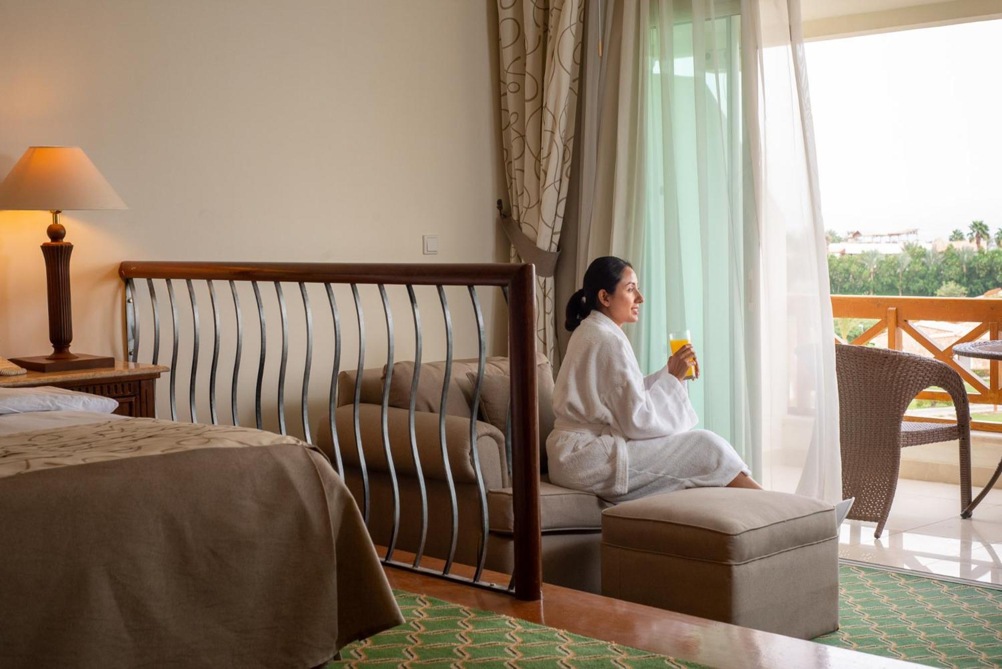 Movenpick Taba Resort & Spa Eksteriør billede The photo shows a cozy hotel room with elegant decor. In the foreground, there is a bed with a neutral-colored blanket and a stylish bed frame with decorative metalwork. A woman in a white bathrobe is sitting on a small ottoman, enjoying a drink. She