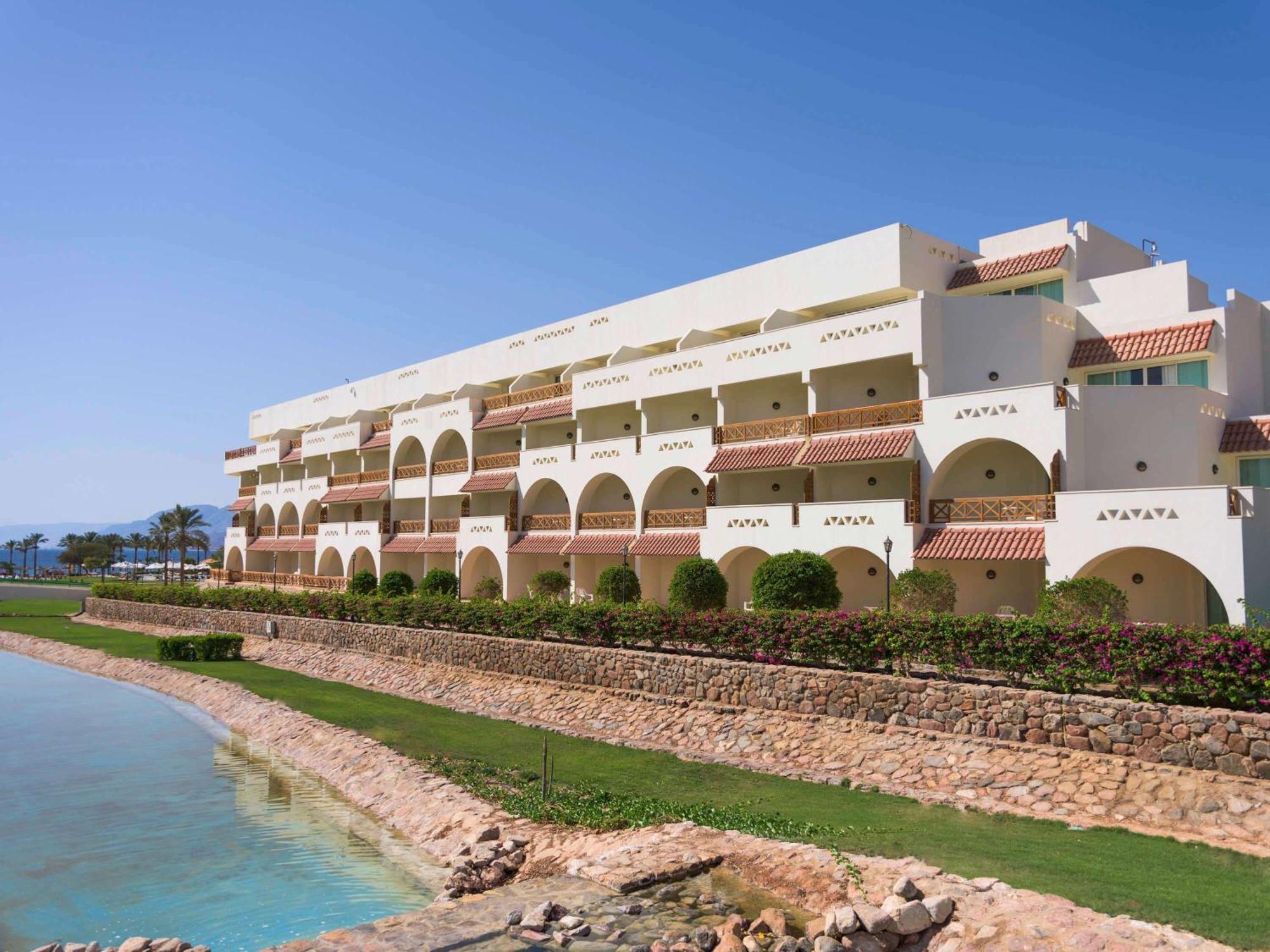 Movenpick Taba Resort & Spa Eksteriør billede The photo shows a modern, multi-story building with a white and light-colored exterior design. It features multiple arches and balconies, giving it a Mediterranean architectural style. The building is situated near a body of water, possibly a lagoon 