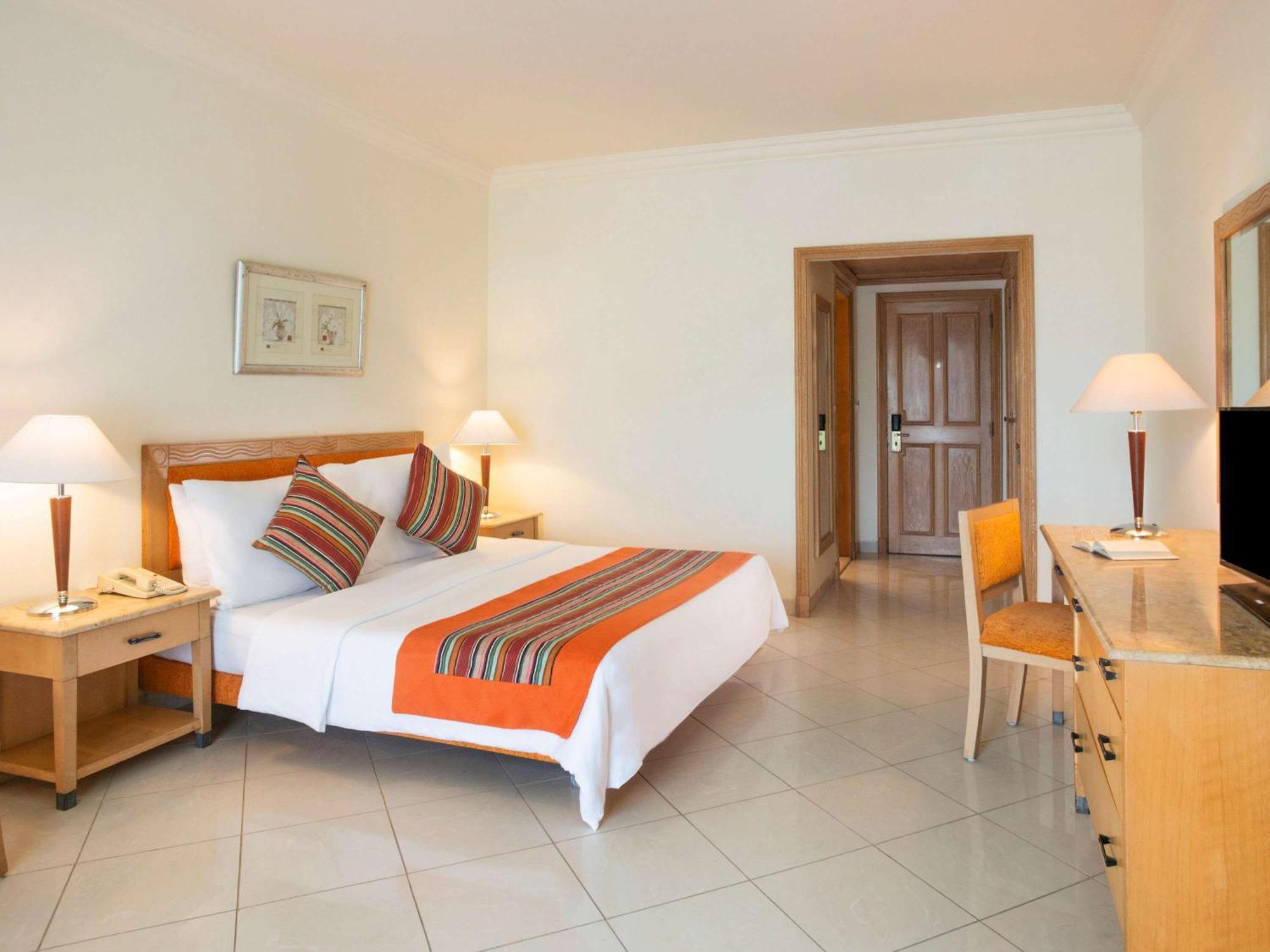 Movenpick Taba Resort & Spa Eksteriør billede The photo shows a well-lit hotel room that features a double bed with a white bedspread and decorative pillows in vibrant colors. There's an orange bed runner across the bed. The room has a simple and modern design, with tiled flooring. 

On one side