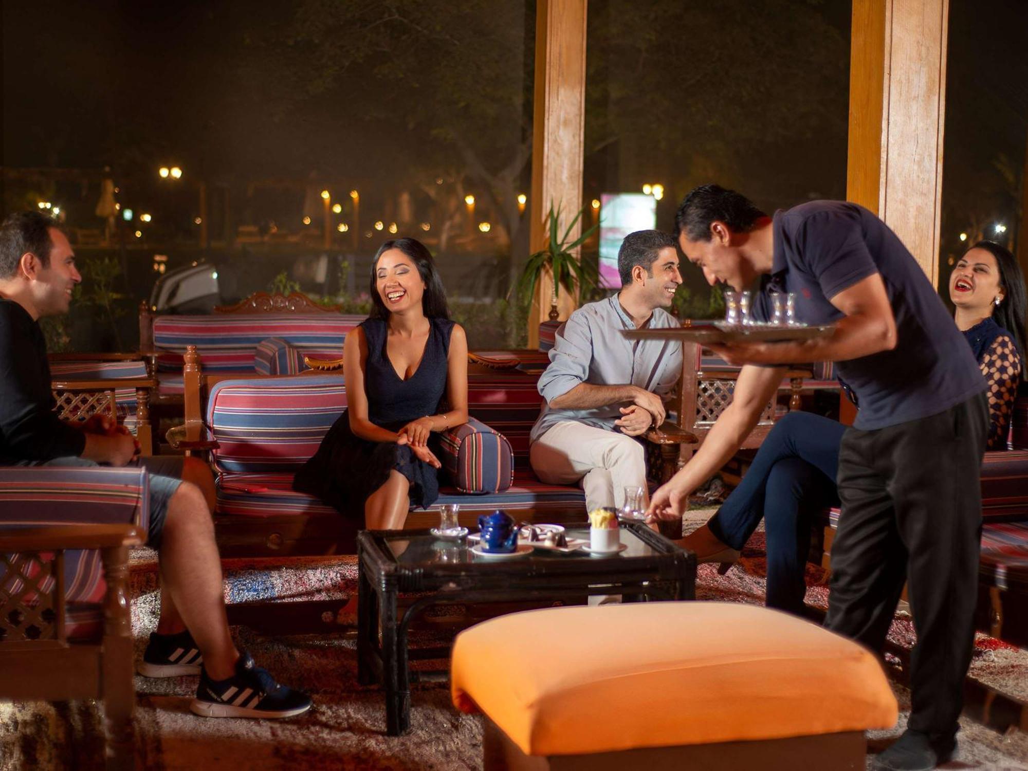 Movenpick Taba Resort & Spa Eksteriør billede The photo depicts a cozy indoor setting with a group of people enjoying each other's company. There are four individuals seated on comfortable furniture, engaged in conversation and laughter. A waiter is serving drinks from a tray to the group. The a