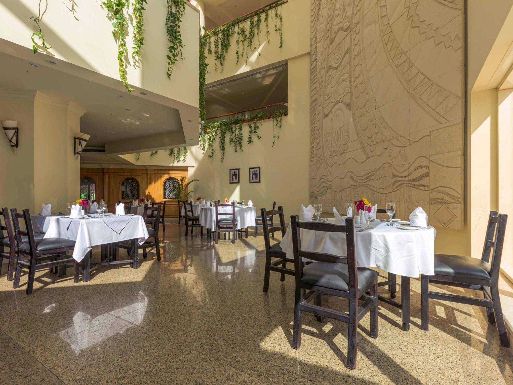 Movenpick Taba Resort & Spa Eksteriør billede The photo shows an indoor dining area of a restaurant. There are several tables, each covered with white tablecloths, and arranged with utensils and flower centerpieces. The space features a high ceiling, with plants hanging down from it, adding a to