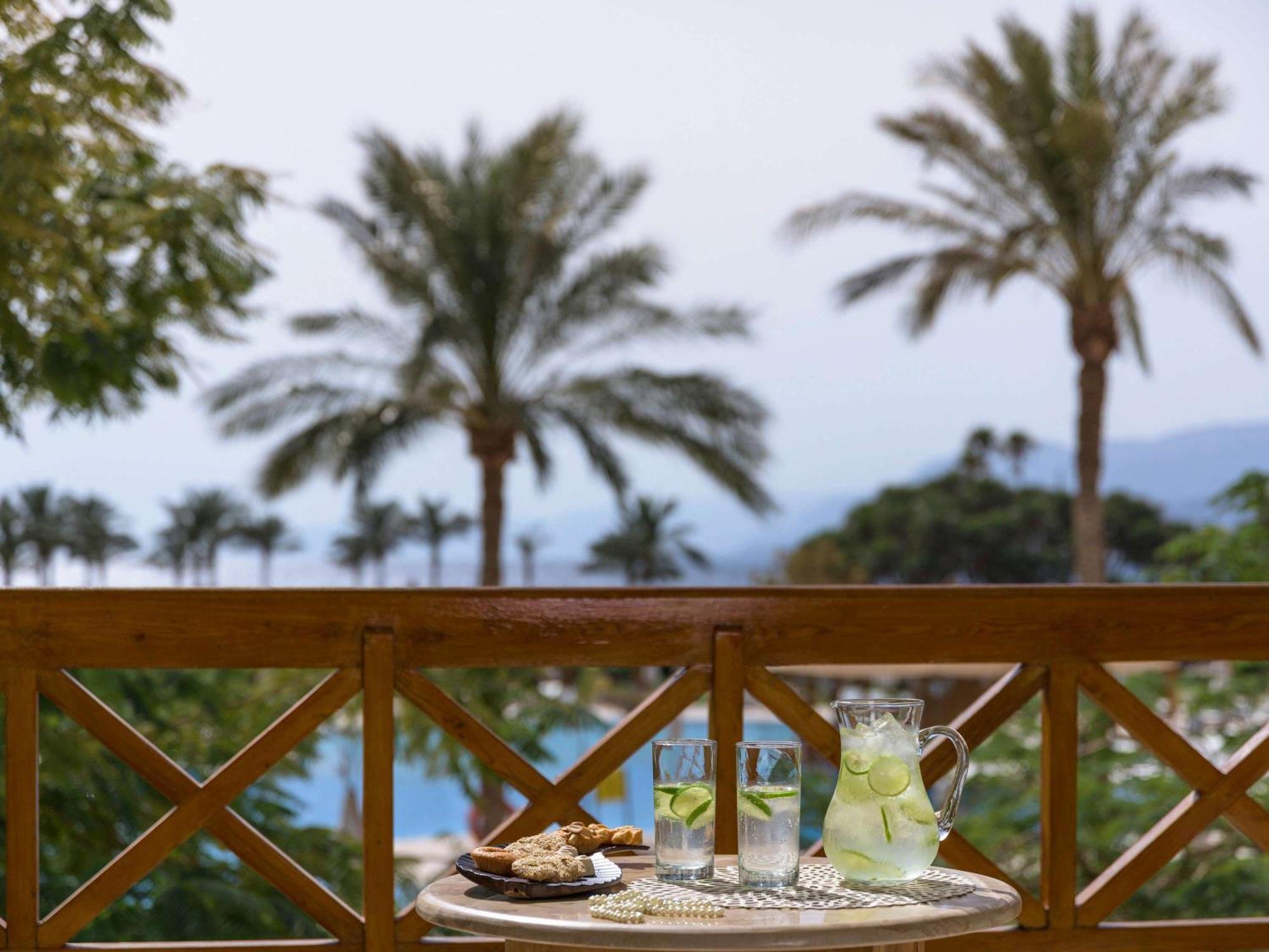 Movenpick Taba Resort & Spa Eksteriør billede The photo shows a scenic outdoor view with several tall palm trees in the background, indicating a tropical or warm environment. In the foreground, there's a small table with a tray that has two clear glasses filled with a light beverage, likely lemo