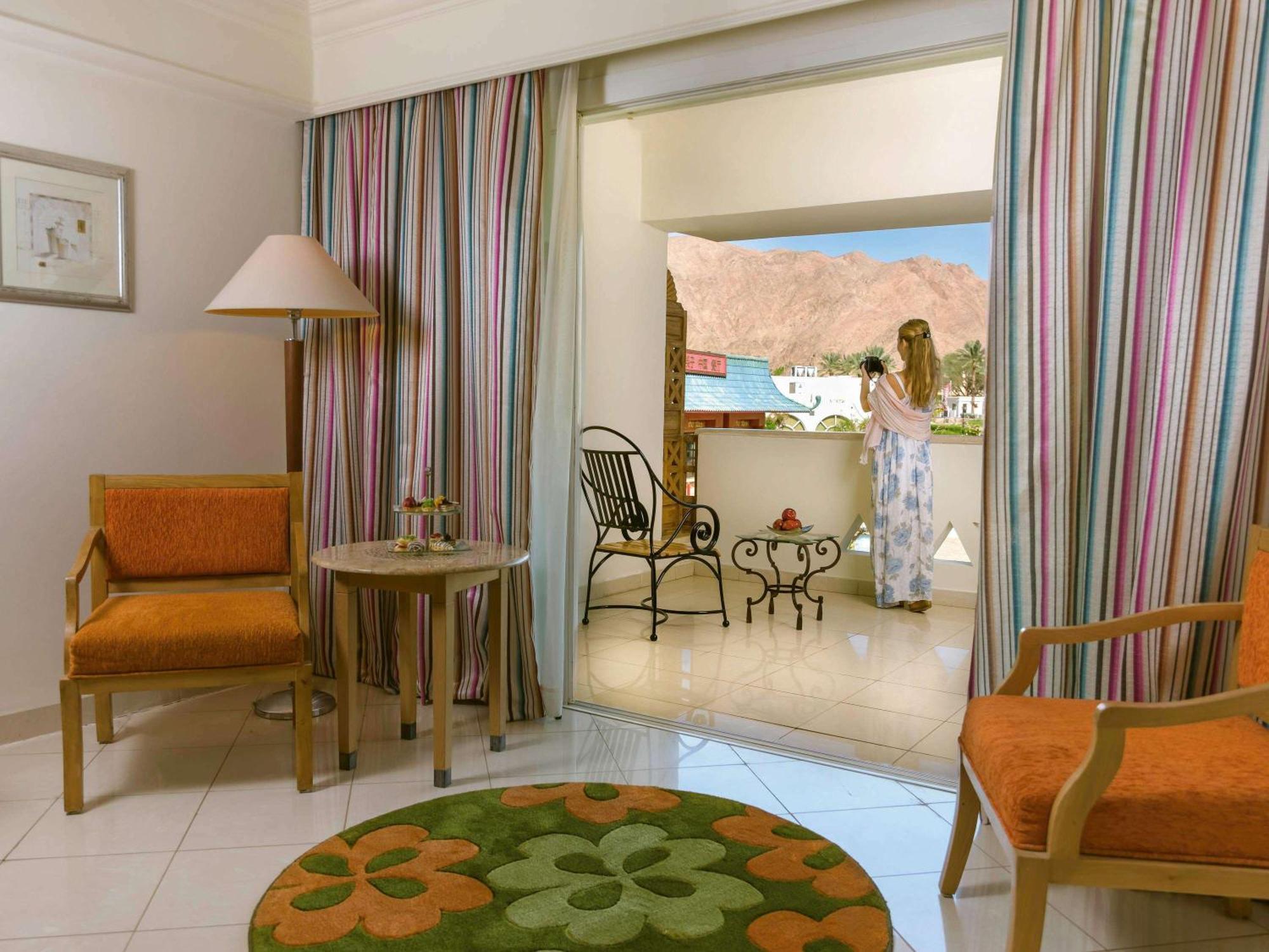 Movenpick Taba Resort & Spa Eksteriør billede The image shows a room with a cozy and inviting atmosphere. It features two orange chairs with wooden frames positioned around a small round table. There is a decorative rug on the floor with a floral pattern in green and orange. 

In the background,