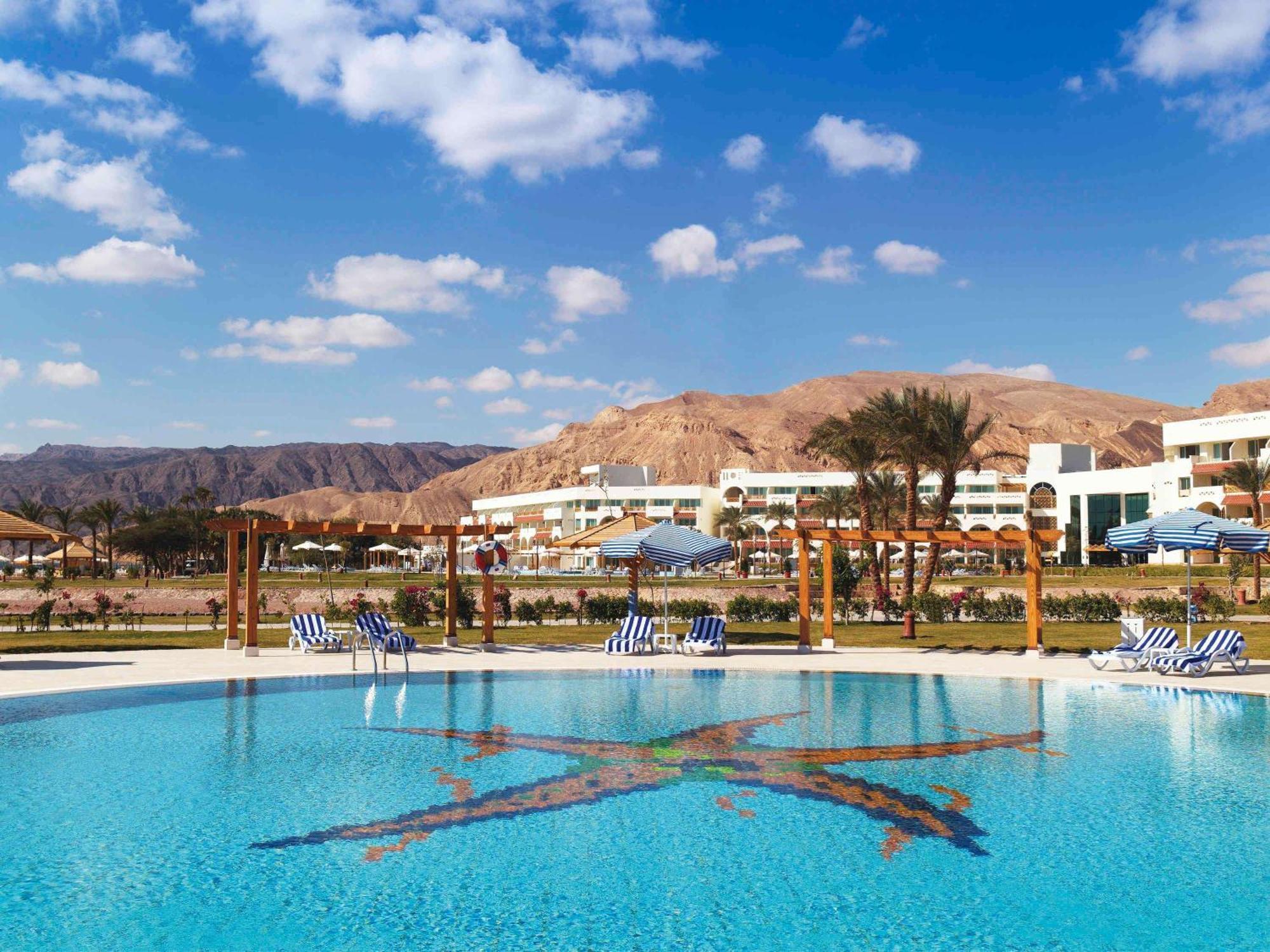 Movenpick Taba Resort & Spa Eksteriør billede The photo shows a tranquil swimming pool set against a backdrop of mountains. The pool's water is clear and reflects a beautiful blue sky with some fluffy clouds. Surrounding the pool are several lounge chairs, and the area appears to be part of a re