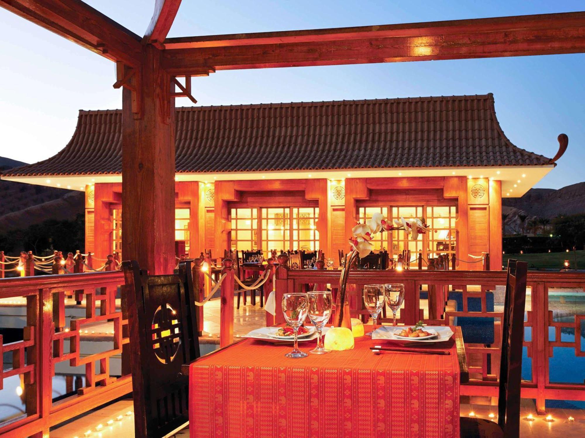 Movenpick Taba Resort & Spa Eksteriør billede The photo shows an outdoor dining setting at a beautifully designed restaurant with traditional architecture. The structure has a distinctive roof and is warmly illuminated, creating a cozy atmosphere at dusk. In the foreground, there is a table set 