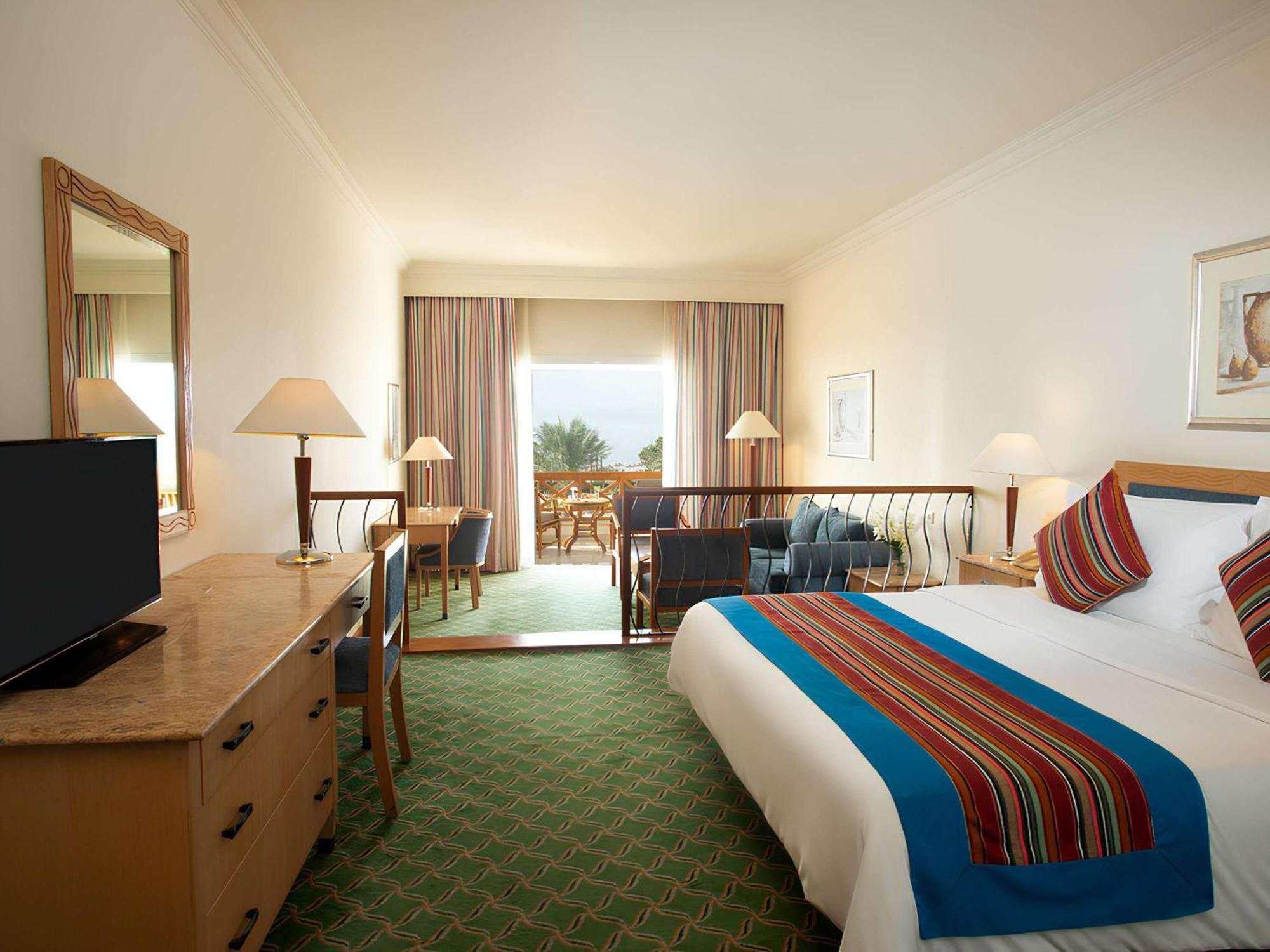 Movenpick Taba Resort & Spa Eksteriør billede The photograph shows a hotel room with a warm and inviting atmosphere. It features a large bed covered with a colorful blanket, which adds a touch of vibrancy to the space. There are two bedside lamps on either side of the bed, providing soft lightin