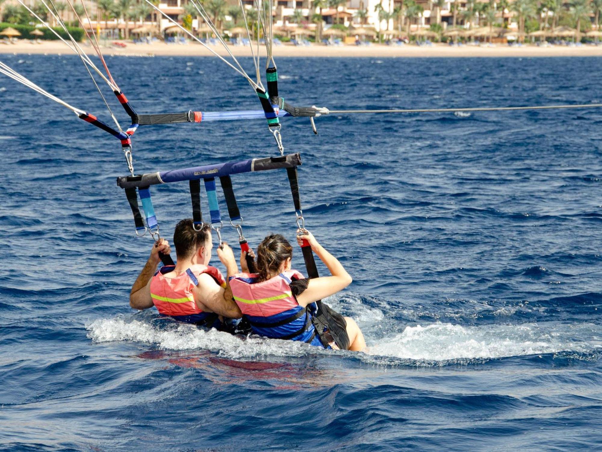 Movenpick Taba Resort & Spa Eksteriør billede The photo shows two people participating in a water sport activity, likely parasailing. They are seated side by side in a harness that is connected to a parachute being towed by a boat. Both individuals are wearing life jackets for safety. The settin