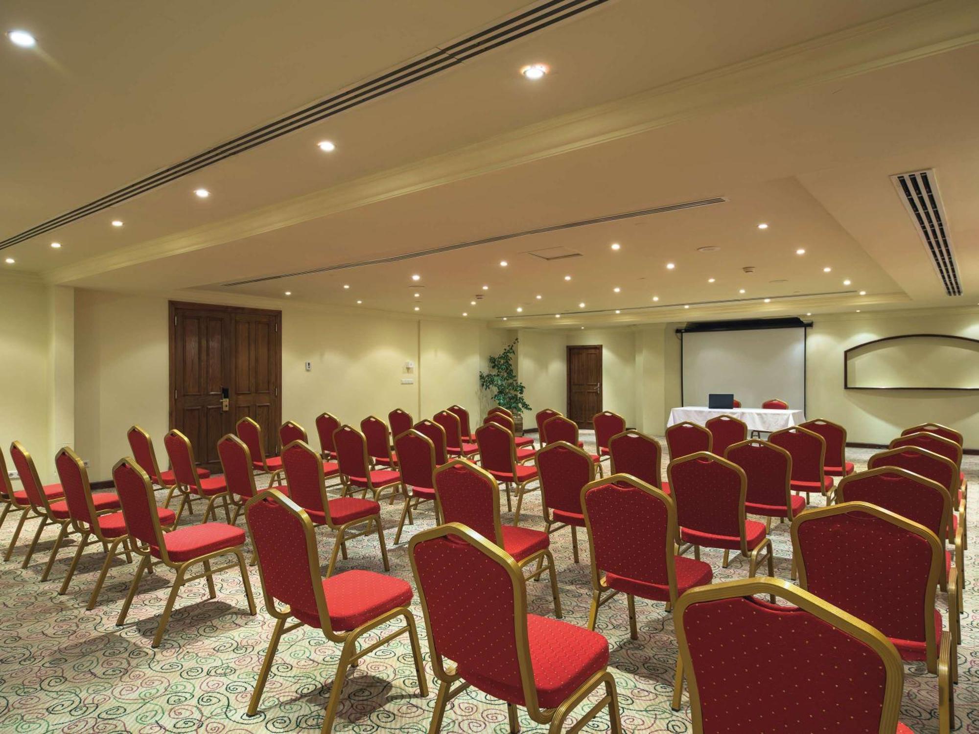 Movenpick Taba Resort & Spa Eksteriør billede The photo shows a conference room set up for a meeting or presentation. There are rows of red chairs arranged facing a front area where a presentation might take place. The room has neutral-colored walls and a carpet with a patterned design. Lighting