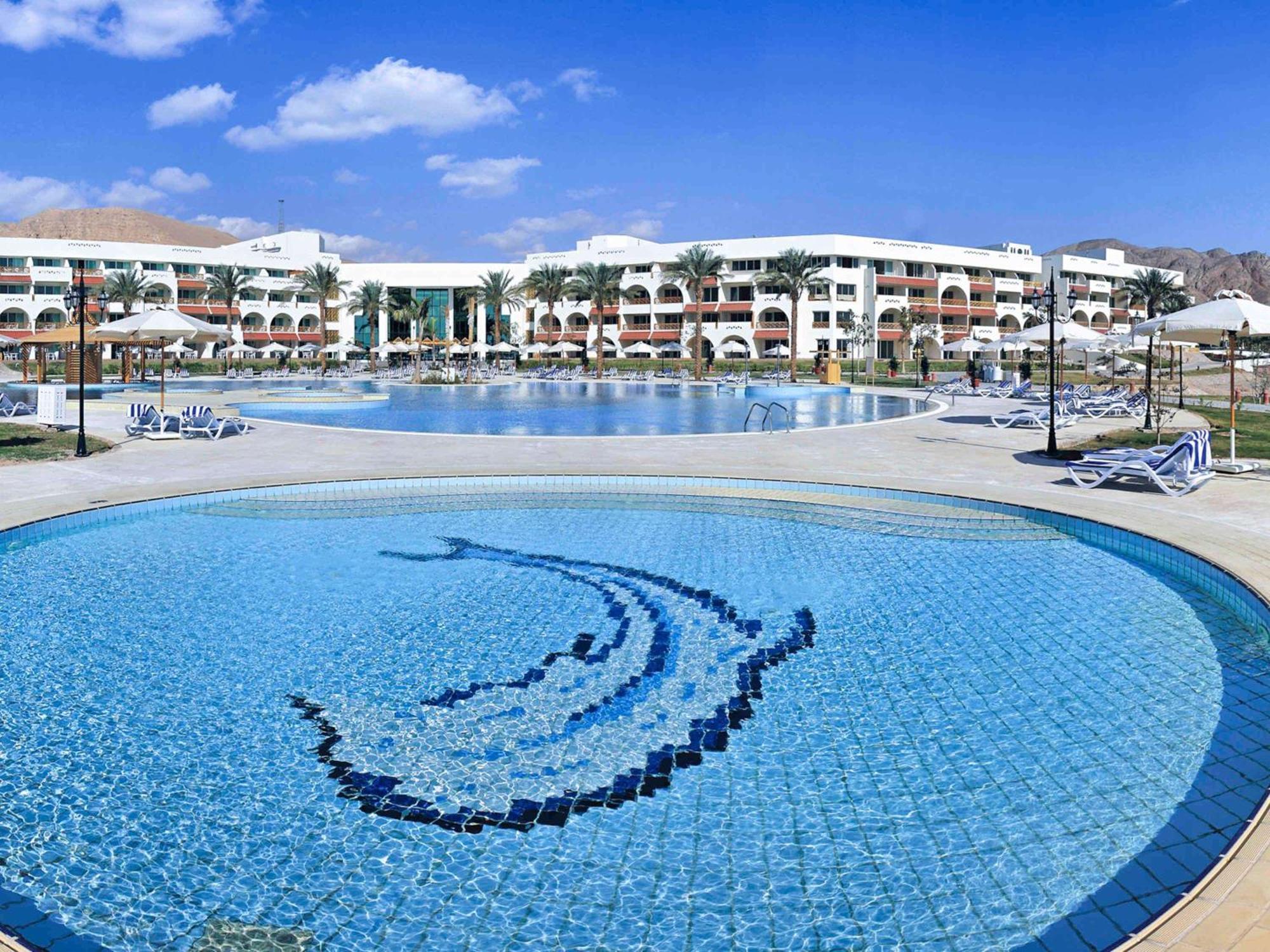 Movenpick Taba Resort & Spa Eksteriør billede The photo shows a beautiful resort area featuring a large swimming pool surrounded by palm trees and lounge chairs. In the foreground, there is a circular mosaic pool with a dolphin design. The background reveals multiple buildings of the resort, set
