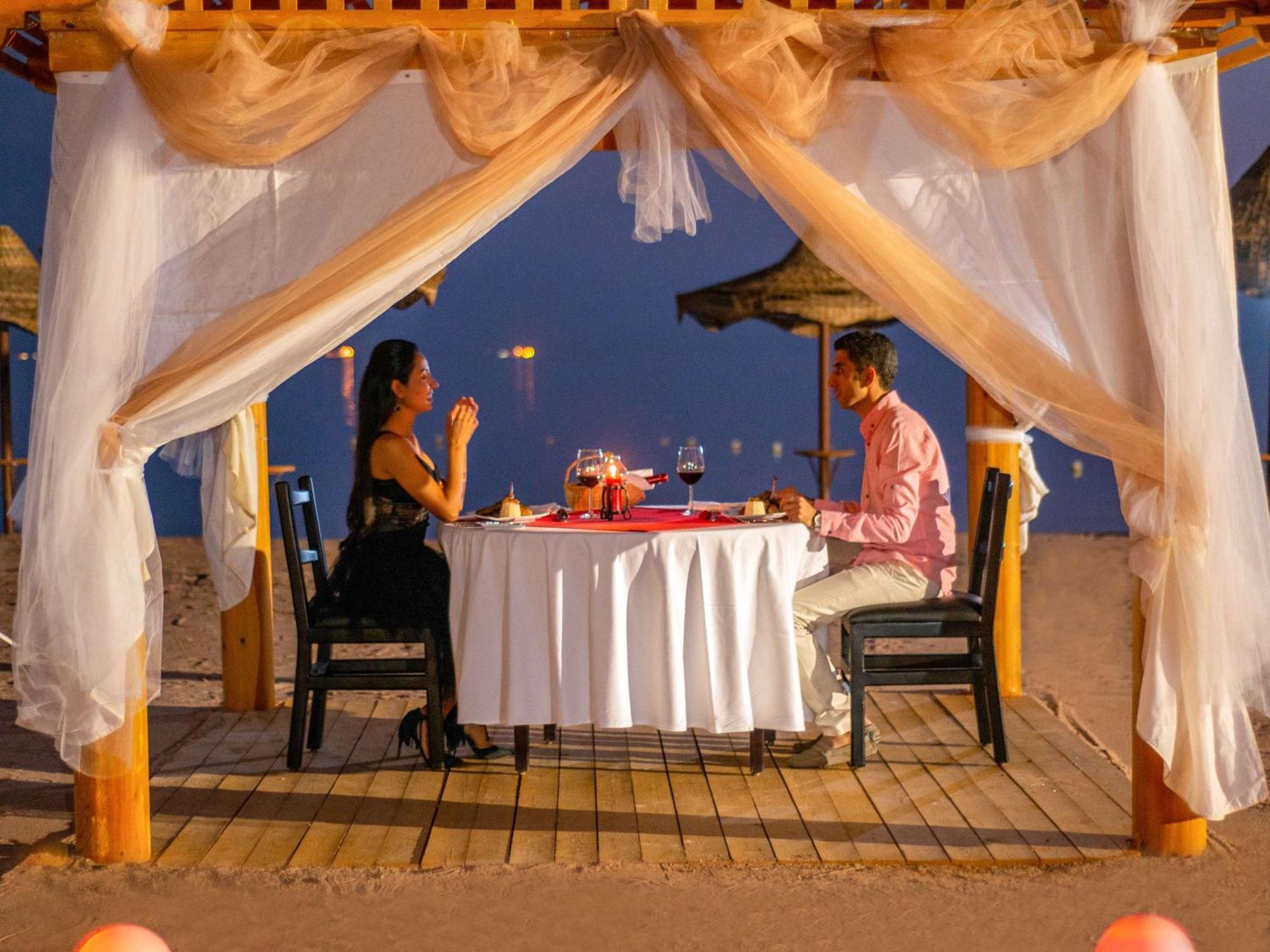 Movenpick Taba Resort & Spa Eksteriør billede The photo depicts a romantic dining setup on the beach. A table is elegantly set with a white tablecloth and decor, including wine glasses and a centerpiece. There are two people seated at the table: one in a black dress and the other in a pink shirt