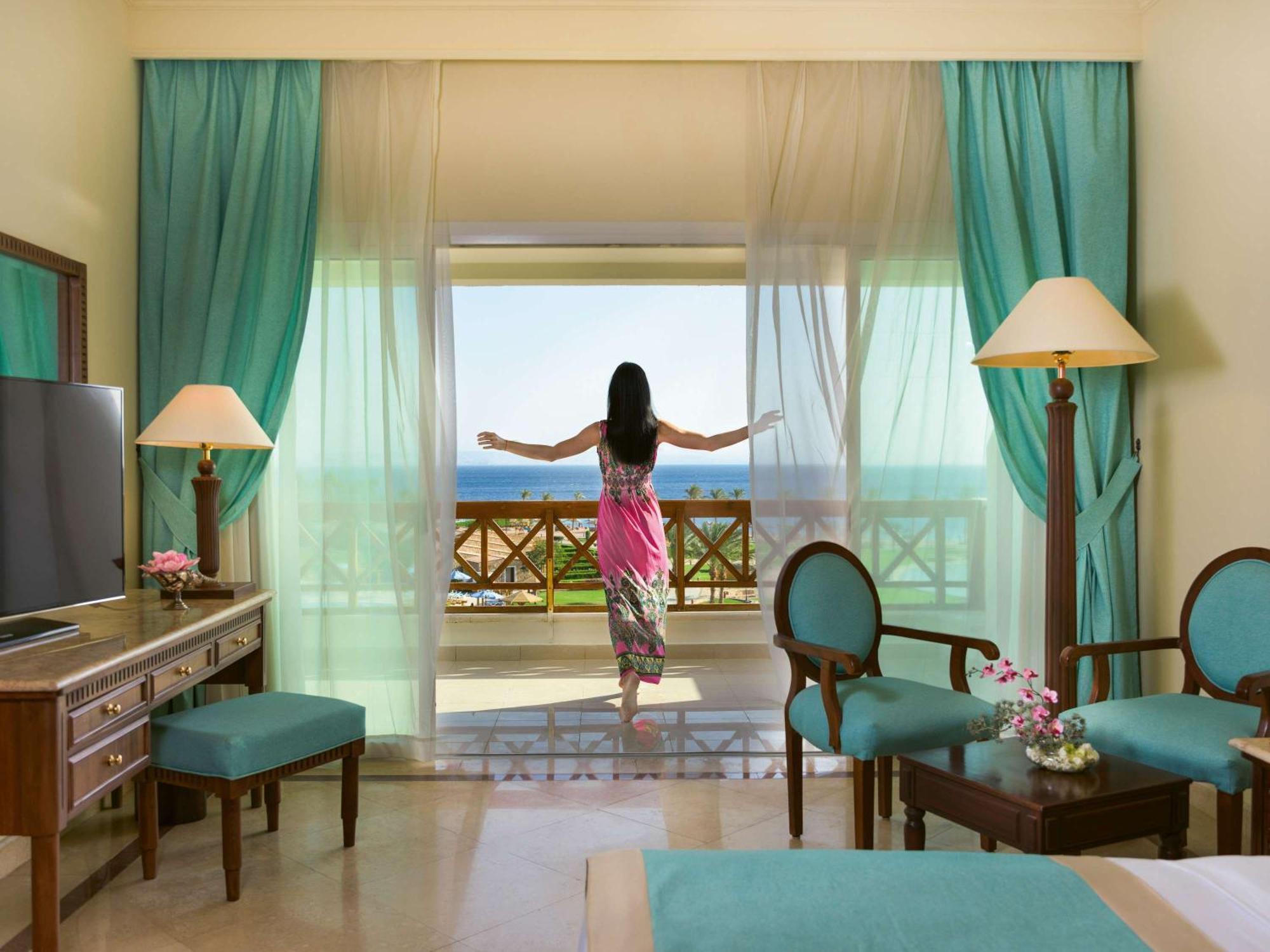 Movenpick Taba Resort & Spa Eksteriør billede The photo shows a serene hotel room with a person standing by a large glass door, enjoying a beautiful view. The room features light-colored walls, turquoise curtains, and elegant furniture, including a TV, a wooden desk, and chairs with matching uph
