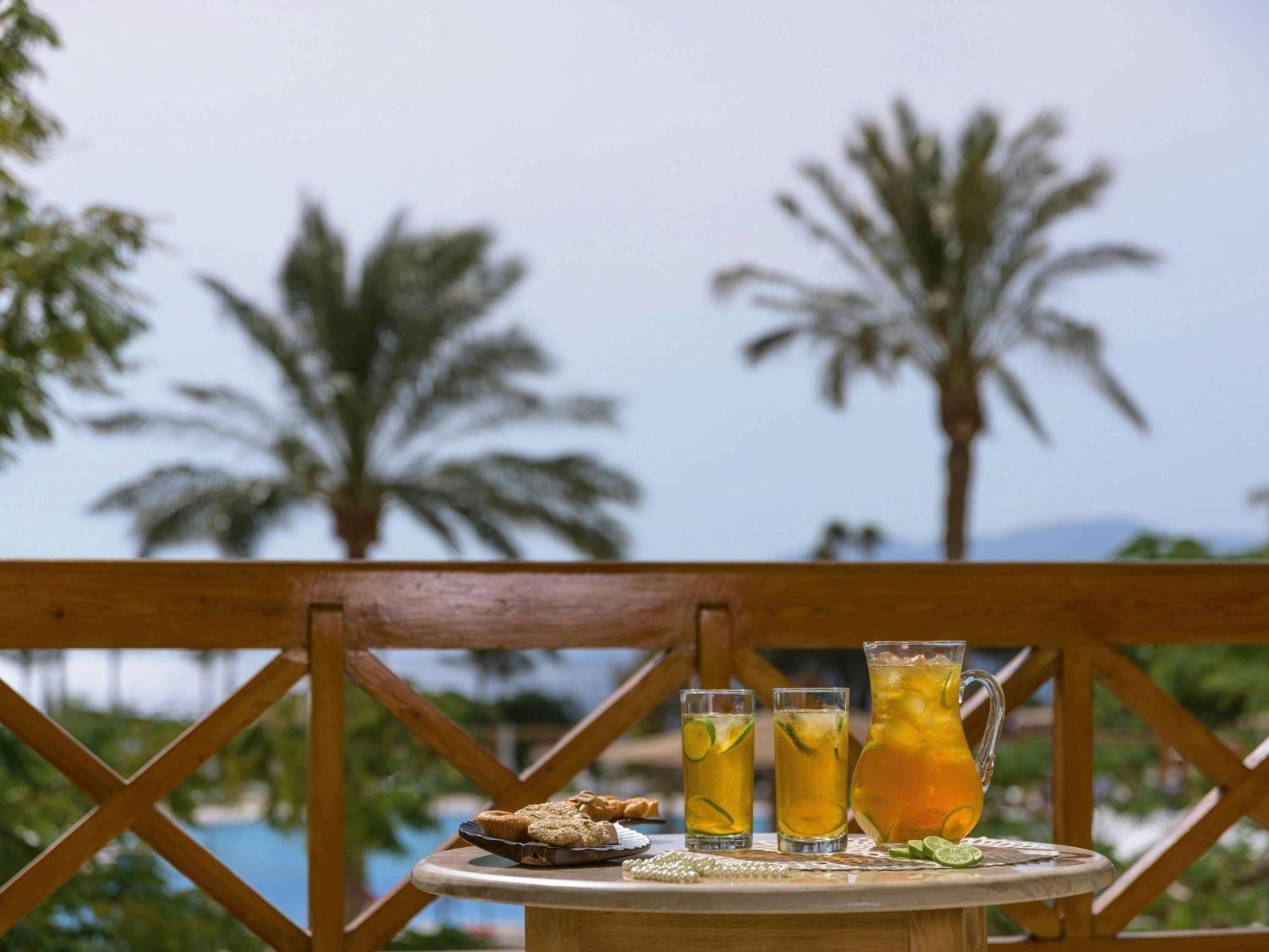 Movenpick Taba Resort & Spa Eksteriør billede The photo shows a table set on a balcony with a view of palm trees in the background. On the table, there are three glasses filled with a yellow drink, likely iced tea or lemonade, garnished with lemon slices. Next to the glasses, there is a pitcher 