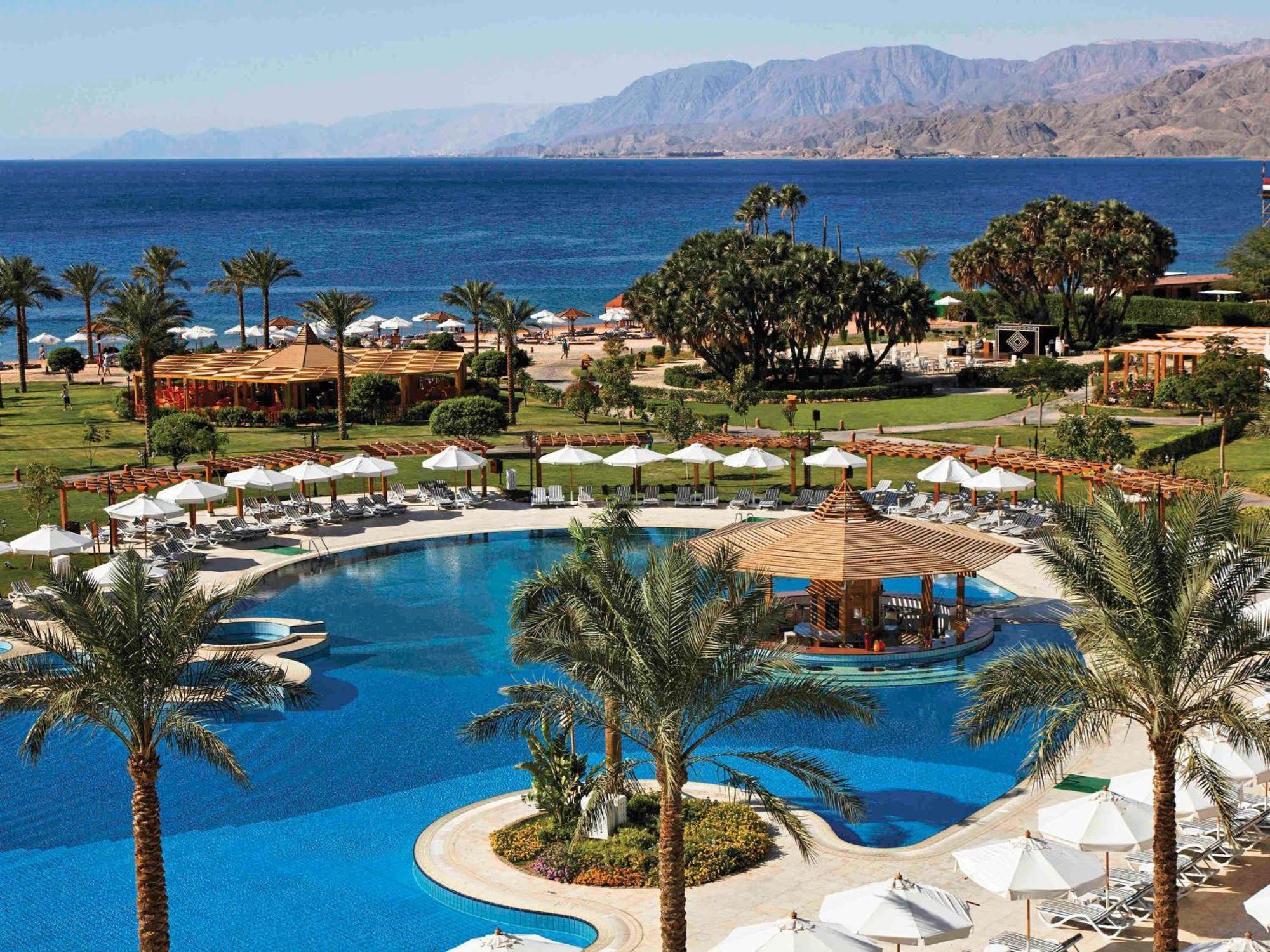 Movenpick Taba Resort & Spa Eksteriør billede The photo shows a luxurious resort overlooking a large, blue swimming pool. Surrounding the pool are palm trees and lounge chairs with white umbrellas, creating a relaxing atmosphere. In the background, the ocean can be seen, with a mountainous lands
